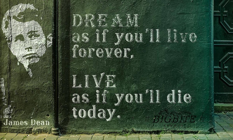 May include: A green wall with white text that reads "DREAM as if you'll live forever, LIVE as if you'll die today." The text is in a vintage font style. The quote is attributed to James Dean. The text is on a green wall with a black metal gate in the background.