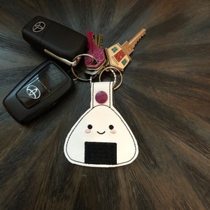 May include: White onigiri keychain with a black filling and a pink button. The keychain has a happy face embroidered on it. The keychain is attached to a set of keys with a house key, a car key, and a small pink charm.
