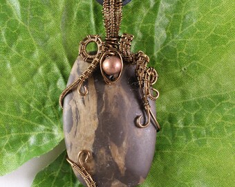 May include: A handcrafted pendant featuring a polished, oval-shaped stone with brown and tan patterns. The stone is intricately wrapped with bronze-colored wire, and a small copper bead is incorporated into the design. The pendant hangs from a black cord.