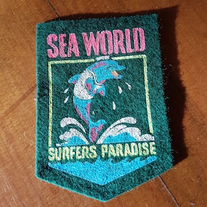 Op de afbeelding: Groen vilt patch met een witte rand en de tekst "SEA WORLD SURFERS PARADISE" in gele en roze letters. Een blauwe en roze dolfijn springt uit het water, dat in wit en blauw is afgebeeld.