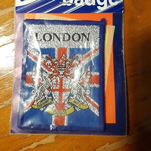 May include: A blue embroidered badge with the text "LONDON" in white lettering. The badge features a silver embroidered Union Jack flag with a silver coat of arms in the center. The coat of arms has a silver cross and two silver lions with gold crowns. The text "DOMINE" and "DIRIGE" is written on a gold banner below the coat of arms.