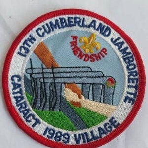 Peut inclure: Un écusson brodé rouge, blanc et bleu avec le texte "13th Cumberland Jamboree Cataract 1999 Village" et un graphique d'un pont avec le mot "Friendship" au-dessus.