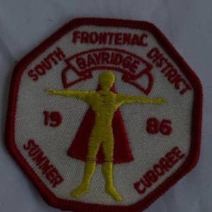 May include: A red and white octagonal patch with the text "South Frontenac District Bayridge" and "1986" in white. The patch also features a yellow stick figure with a red cape.