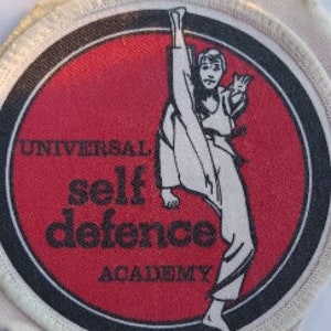 Puede incluir: Un parche redondo blanco con borde negro y centro rojo. El parche presenta una ilustración en blanco y negro de una persona en una pose de artes marciales. El texto "Universal self defence academy" está impreso en negro sobre fondo rojo.