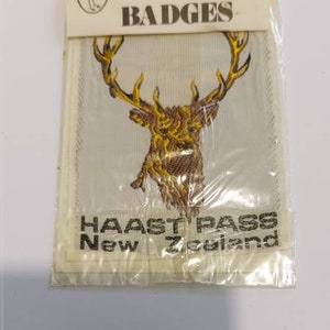 May include: A white and gold embroidered patch featuring a stag's head and the text "HAAST PASS New Zealand". The patch is packaged in a clear plastic bag with a white label that reads "ROTURU White Heron of N.Z. BADGES".