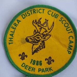 May include: A circular, yellow Cub Scout camp badge with a green border. It displays a deer head, the text "THALERA DISTRICT CUB SCOUT CAMP", the year "1986", and the words "DEER PARK".
