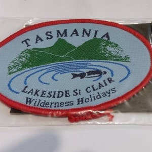 May include: A red, white, and blue embroidered patch with the text "TASMANIA LAKESIDE St CLAIR Wilderness Holidays" and a depiction of a lake and mountains. The patch is attached to a black card with the text "WOMBAT BADGE $2.95 AUSTRALIA".