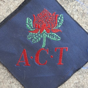May include: A dark blue diamond-shaped patch with red embroidered lettering that reads "A.C.T." and a red and green embroidered flower in the center.