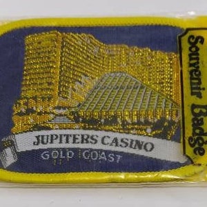 Puede incluir: Una insignia de recuerdo amarilla y azul con el texto "Jupiters Casino Gold Coast" y "Souvenir Badge" sobre un fondo blanco. La insignia presenta una imagen dorada y plateada de un edificio.