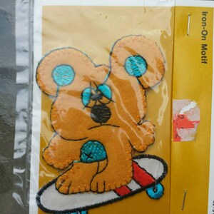 May include: Iron-on motif featuring a brown bear wearing a blue hat and riding a skateboard with red and white stripes. The bear has blue eyes and a black nose. The skateboard has blue wheels.