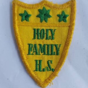 Könnte beinhalten: Gelber Filz-Aufnäher mit einem grün gestickten Schild. Der Schild hat oben drei grüne Sterne und den Text "HOLY FAMILY H.S." in grün.