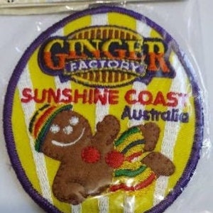 May include: A round, embroidered patch with a purple border. The centre features a yellow and white striped background with a gingerbread man in the centre. The gingerbread man is brown with a white face and red cheeks. The text "Ginger Factory Sunshine Coast Australia" is printed in a yellow, orange, and red font.