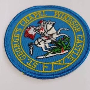 Könnte beinhalten: Ein blauer und gelber gestickter Aufnäher mit dem Text "St. George's Chapel Windsor Castle" und einer Darstellung eines Ritters auf einem Pferd, der einen Drachen tötet.