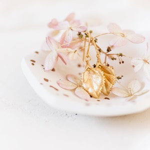 Pu&ograve; includere: Orecchini a foglia d'oro con un design delicato, esposti su un piatto bianco con petali di fiori rosa secchi.