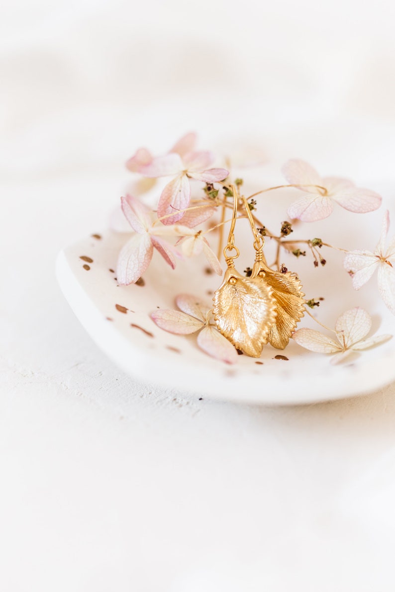 Pu&ograve; includere: Orecchini a foglia d'oro con un design delicato, esposti su un piatto bianco con petali di fiori rosa secchi.
