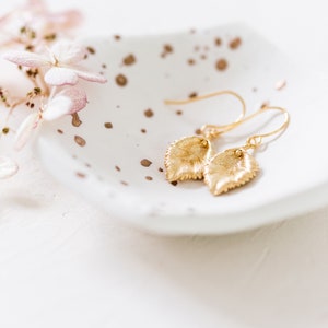 Pu&ograve; includere: Un paio di orecchini a foglia d'oro su un piatto bianco macchiato con un fiore rosa sullo sfondo.