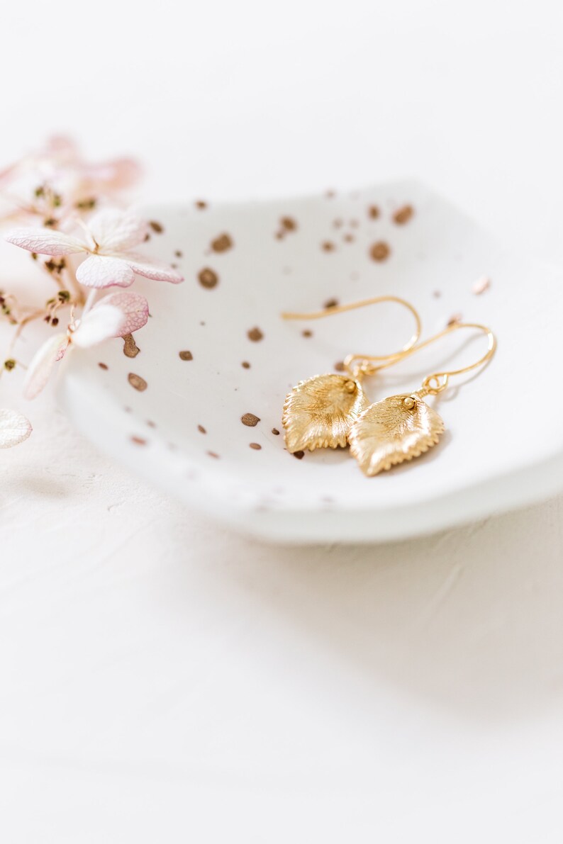 Pu&ograve; includere: Un paio di orecchini a foglia d'oro su un piatto bianco macchiato con un fiore rosa sullo sfondo.
