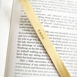K&ouml;nnte beinhalten: Ein langes, d&uuml;nnes Lesezeichen aus hellem Holz mit einer geschwungenen Oberkante, das in einem offenen Buch liegt. Der Name Emma ist in einer Serifenschrift vertikal in der Mitte des Lesezeichens eingraviert, das &uuml;ber dem Text liegt.