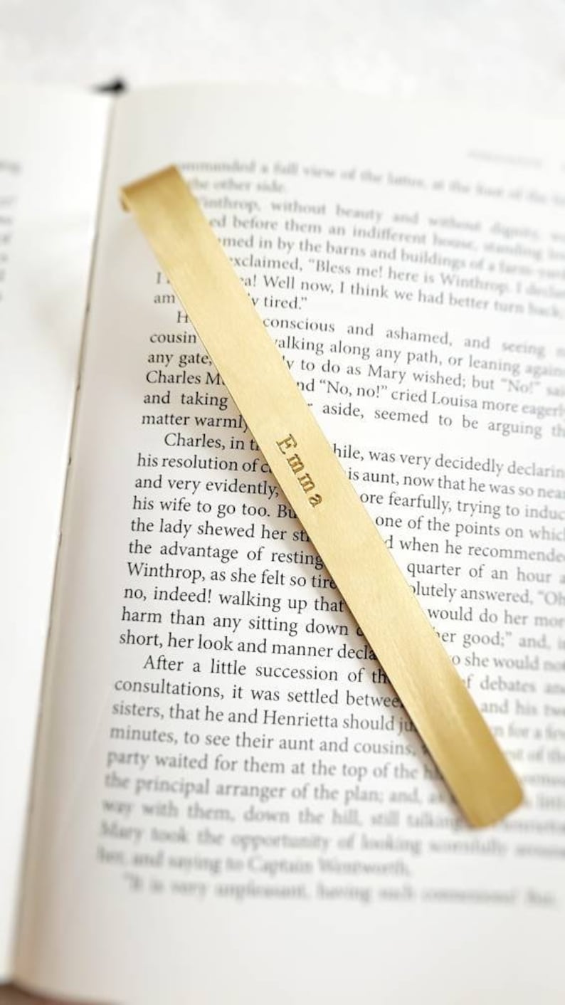 K&ouml;nnte beinhalten: Ein langes, d&uuml;nnes Lesezeichen aus hellem Holz mit einer geschwungenen Oberkante, das in einem offenen Buch liegt. Der Name Emma ist in einer Serifenschrift vertikal in der Mitte des Lesezeichens eingraviert, das &uuml;ber dem Text liegt.