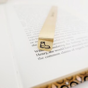 K&ouml;nnte beinhalten: Ein goldfarbenes Lesezeichen aus Metall mit einem rechteckigen Ende, das eine schwarze Gravur eines Schn&uuml;rstiefels aufweist. Das Lesezeichen ist vertikal auf einem aufgeschlagenen Buch platziert, wobei gedruckter Text auf den wei&szlig;en Seiten sichtbar ist.