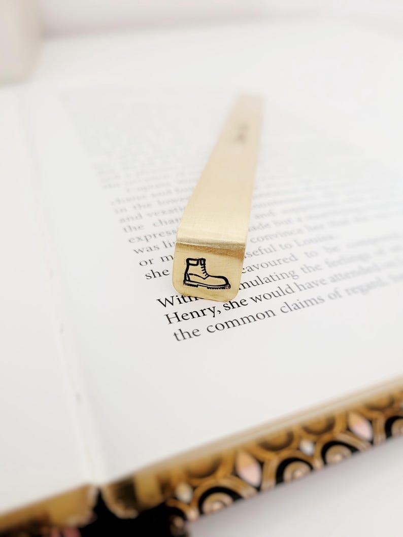 K&ouml;nnte beinhalten: Ein goldfarbenes Lesezeichen aus Metall mit einem rechteckigen Ende, das eine schwarze Gravur eines Schn&uuml;rstiefels aufweist. Das Lesezeichen ist vertikal auf einem aufgeschlagenen Buch platziert, wobei gedruckter Text auf den wei&szlig;en Seiten sichtbar ist.