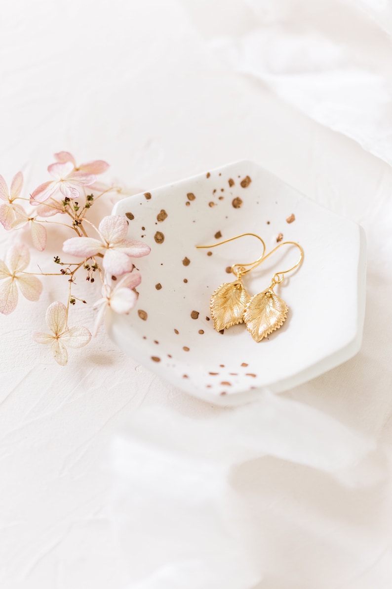 Pu&ograve; includere: Orecchini a foglia d'oro a forma di foglie su un piatto bianco con macchie dorate.