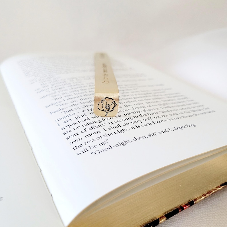 K&ouml;nnte beinhalten: Ein rechteckiges Lesezeichen aus goldfarbenem Metall, das auf einer offenen Buchseite liegt. Das Lesezeichen zeigt am unteren Ende eine schwarze, eingravierte Strichzeichnung einer Mohnblume sowie vertikal eingravierten Text entlang der L&auml;nge.
