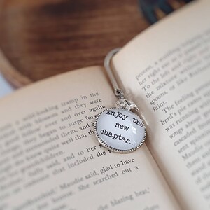 May include: A silver-toned metal bookmark featuring a circular charm with a white background and the black text Enjoy the new chapter. The charm is attached to a curved metal hook designed to slide between the pages of an open book.
