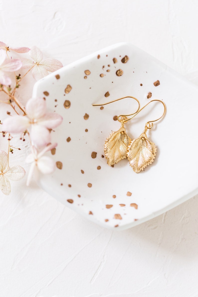 Pu&ograve; includere: Un paio di orecchini a foglia d'oro su un piatto bianco con macchie dorate. Gli orecchini hanno la forma di foglie e un delicato design a filo.