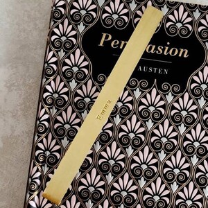 K&ouml;nnte beinhalten: Ein goldfarbenes Lesezeichen aus Metall mit der eingravierten Aufschrift Emma, das diagonal auf einem gebundenen Buch mit dem Titel Persuasion von Austen liegt. Der Einband zeigt ein sich wiederholendes schwarz-rosa-silbernes F&auml;chermuster.