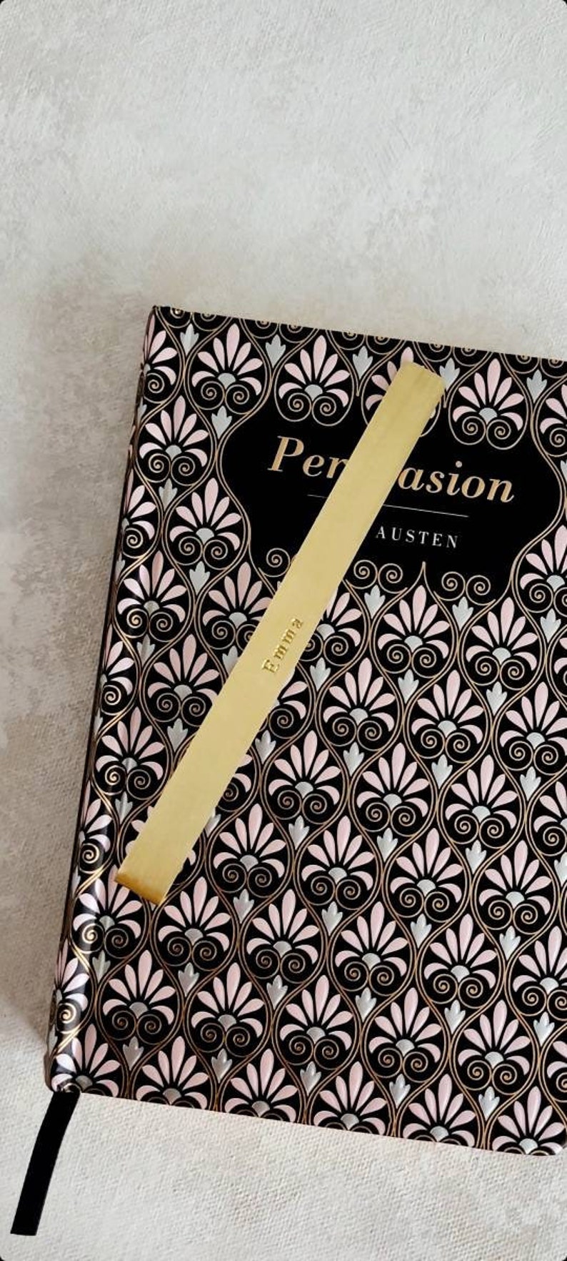 K&ouml;nnte beinhalten: Ein goldfarbenes Lesezeichen aus Metall mit der eingravierten Aufschrift Emma, das diagonal auf einem gebundenen Buch mit dem Titel Persuasion von Austen liegt. Der Einband zeigt ein sich wiederholendes schwarz-rosa-silbernes F&auml;chermuster.