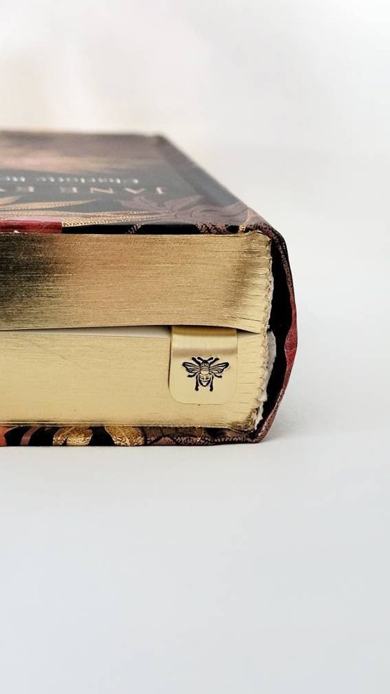 K&ouml;nnte beinhalten: Ein gebundenes Buch mit goldfarbenen Seitenschnitten und einem dunklen Einband mit Blumenmuster. Ein kleiner, goldfarbener Lesezeichen-Clip aus Metall mit einer schwarzen Bienenillustration ist am unteren Seitenrand befestigt.