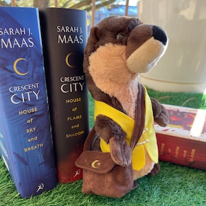 May include: A plush otter toy wearing a yellow vest and a brown satchel with a gold crescent moon symbol, positioned next to two books titled Crescent City: House of Sky and Breath and Crescent City: House of Flame and Shadow by Sarah J. Maas.