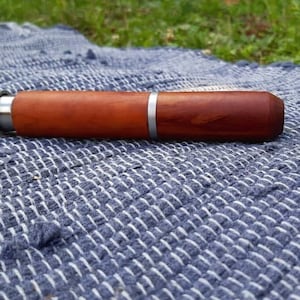 Könnte beinhalten: Ein Holz- und Metallstift mit einem silbernen Band. Der Stift liegt auf einem blau-weiß gestreiften Teppich.