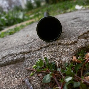 May include: A black metal pipe lying on a grey concrete surface with green plants growing around it.