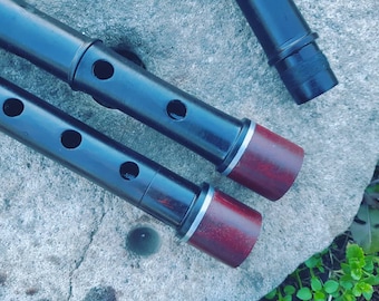 May include: Close-up of several black wooden flutes with dark red ends and silver bands. The flutes have multiple finger holes and are arranged on a gray stone surface. The image highlights the craftsmanship of the musical instruments.