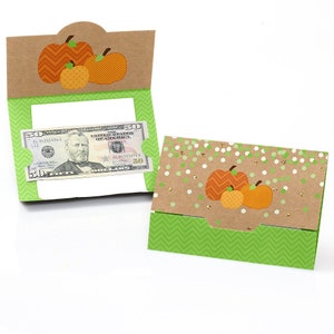 May include: A set of three brown and orange pumpkin-shaped gift card holders with a green and white chevron pattern on the bottom. The holders are made of cardstock and have a slot for inserting a gift card or cash. The holders are decorated with a confetti pattern and a gold glitter accent.