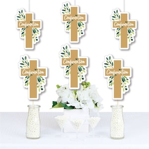 May include: Six green and gold cross-shaped table decorations with the word "Confirmation" on each. The decorations are on sticks and are displayed in a table setting with white flowers and white candy.