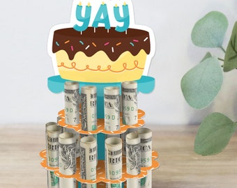 May include: A multi-tiered birthday cake stand with a blue and yellow cake topper that says "YAY". The stand is decorated with orange and white flowers and holds rolled up dollar bills.