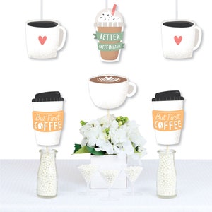 May include: Coffee-themed party decorations featuring paper cutouts of coffee cups, a latte, and a to-go coffee cup with the text "But First, Coffee." The decorations include a white floral arrangement and martini glasses filled with white beads.