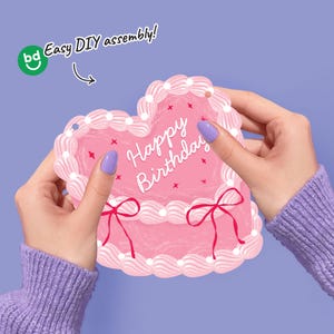 May include: A pink heart-shaped birthday card with white frosting details and red bows. The card has the words "Happy Birthday" written in white. The card is held by two hands, and the text "Easy DIY assembly!" is visible.