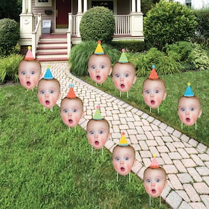 May include: Nine colorful cardboard cutouts of baby faces with party hats. The cutouts are placed on a green lawn with a brick walkway.