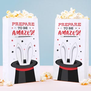 May include: Two white paper popcorn bags with the text "PREPARE TO BE AMAZED!" in red. Each bag features a black top hat with white rabbit ears and is filled with popcorn. Stars and a magic wand are also on the bags.