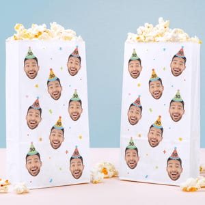 May include: Two white paper popcorn bags filled with popcorn. Each bag is decorated with a repeating pattern of a smiling face wearing a party hat with the number "30" on it. Colorful confetti and stars are scattered around the faces.