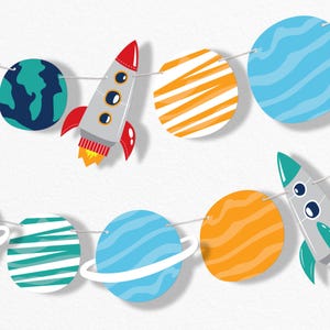 May include: A space-themed banner featuring colorful planets and rockets. The planets are blue, orange, and green with white accents. The rockets are gray with red and yellow details. The banner is strung on a white string.