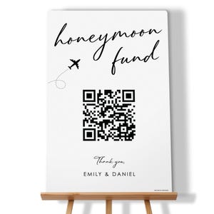 May include: A white sign with the words "honeymoon fund" in elegant script, a small airplane graphic, and a QR code. The sign also includes the text "Thank you, EMILY & DANIEL". The sign is displayed on a wooden easel.