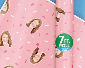 May include: Pink wrapping paper with a repeating pattern of a girl's face and confetti. The paper is rolled up and has a label that says "7 ft. Roll Made in the USA".