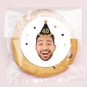 May include: A large chocolate chip cookie in a clear cellophane bag. The cookie has a white circular sticker with a man's face wearing a black and gold party hat with the number 40. Black and gold star accents surround the image.