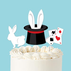 May include: A white frosted cake decorated with a black top hat, white rabbit ears, a white rabbit, and playing cards. The top hat has a red band. The playing cards show the suits of clubs, hearts, and diamonds. The background is light blue.