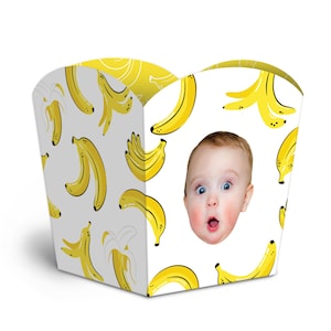 May include: A white paper treat box featuring a repeating pattern of yellow bananas and a centered photographic print of a baby's face with a surprised expression. The container has a curved top edge and a wide, open-top design for holding items.